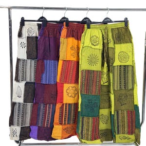 May include: Four pairs of patchwork pants in various colors, including purple, orange, and green, hang on a rolling rack. Each pair features a unique design with floral and geometric patterns. The pants have a drawstring waist.