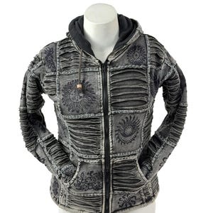 Peut inclure: Sweat à capuche zippé gris avec un motif patchwork. Le sweat à capuche présente une capuche et des poches noires, avec des motifs floraux et solaires décoratifs. Le tissu a un aspect vieilli et texturé. La fermeture éclair est noire, et le cordon a une perle en bois.