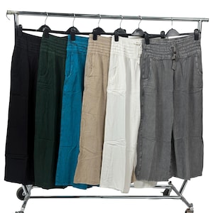 May include: A clothes rail displays wide-leg trousers in various colours: black, dark green, teal, beige, white, and grey. The trousers are made of a lightweight fabric and have a relaxed fit. The rail is on wheels.