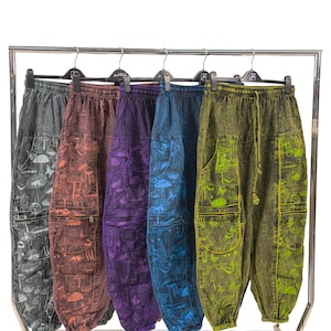 May include: Five pairs of colorful, patterned pants hanging on a metal rack. The pants are in shades of gray, brown, purple, blue, and green. Each pair features a unique mushroom design and a drawstring waist. The pants have pockets on the sides.