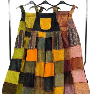 Pode incluir: Três vestidos de patchwork pendurados em um varão de metal. Os vestidos apresentam uma variedade de cores, incluindo verde, laranja, preto e rosa. Cada vestido tem um grande bolso frontal e alças ajustáveis. Os vestidos são feitos de um patchwork de diferentes tecidos.
