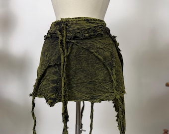 May include: A mini skirt in a dark olive green and black acid-wash denim fabric. The skirt features a high-waisted design with a layered, asymmetrical hemline, raw frayed edges, and multiple long, dangling fabric strips hanging from the waist.