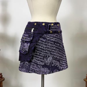 May include: A dark purple mini skirt featuring a white printed pattern of Tibetan script, lions, and seated figures. The skirt includes a side cargo pocket with a zipper closure, a waistband with brass-colored metal buttons, and an adjustable side strap.