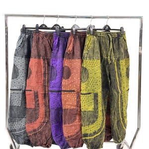 May include: Five pairs of patterned, loose-fitting trousers hang on a clothing rail. The trousers feature geometric designs on the pockets and lower legs. Colours include grey, rust, purple, orange, and olive green.