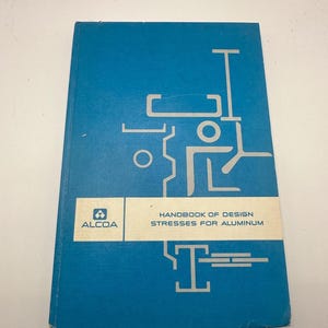 May include: A blue hardcover book titled "Handbook of Design Stresses for Aluminum." The book features a silver geometric design on the cover, with the "ALCOA" logo. The book is a technical manual.