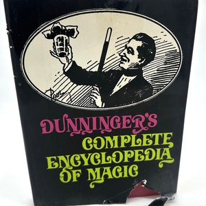 May include: A black book cover with a white oval illustration of a magician holding a wand and a top hat. The book title is "DUNNINGER'S COMPLETE ENCYCLOPEDIA OF MAGIC" in pink and green lettering.