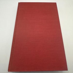 Pode incluir: Um livro de capa dura fechado com uma capa vermelha texturizada. A lombada do livro é visível no lado esquerdo, e a capa parece ser feita de um material tecido. O livro é retangular e está plano sobre uma superfície branca.