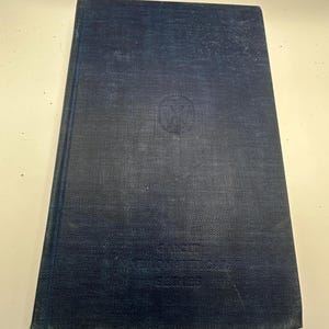 May include: A vintage hardback book with a dark blue cover. The cover has a textured appearance and a circular emblem near the top. The title and series name are embossed near the bottom. The book's edges show signs of wear.