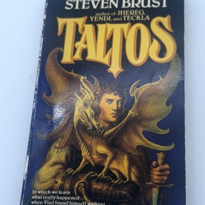 May include: A book cover with a dark blue background featuring a golden dragon and a man holding a dagger. The title of the book is "Taltos" in large yellow letters. The author is Steven Brust, author of "Jhereg, Yendi, and Teckla". The text on the bottom of the cover reads "In which we learn what really happened when Vlad found himself walking the Paths of the Dead."