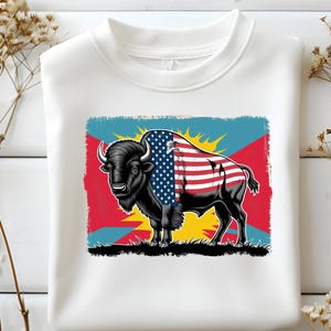 May include: White sweatshirt with a graphic of a black bison, the American flag draped over its back. The bison is set against a colourful background of red, blue, and yellow. The sweatshirt is made of a soft material.