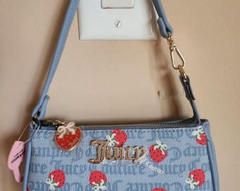 May include: A light blue handbag featuring a strawberry pattern and the word "Juicy" in gold. The bag has a matching blue strap and a gold-coloured clasp. A small tag with the words "Juicy Couture" is attached.