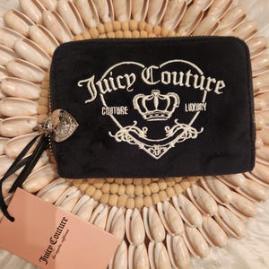 May include: Black Juicy Couture wallet with a white embroidered logo and crown design. The wallet has a silver zipper pull and a black wrist strap. A pink tag with the Juicy Couture logo is attached.