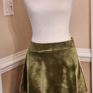 May include: A green velvet mini skirt with a flared hem. The skirt has a small embroidered logo on the bottom right side.