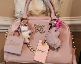 May include: Pale pink Juicy Couture handbag with a gold chain strap and handles. The bag has a large rhinestone-studded logo and several charms, including a small purse and a heart. A tag reads "Juicy Couture".