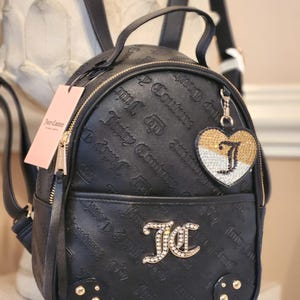 May include: Black backpack with embossed "Juicy Couture" text and gold-tone hardware. Features a heart-shaped charm with the letter "J" and a tag with the brand name. The bag has a zippered closure and adjustable straps.