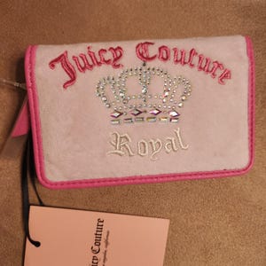 May include: A light pink Juicy Couture wallet with a crown design and the word "Royal". The wallet has a pink border and the brand name is embroidered in pink. A pink tag with the brand name is attached.