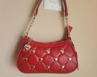 May include: A red quilted handbag featuring a gold-tone chain strap and gold heart-shaped studs embedded in the diamond-patterned exterior. A small heart-shaped charm hangs from the zip pull, and the Juicy Couture logo is centred on the front.