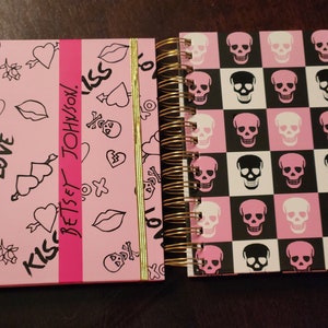 May include: Two spiral-bound notebooks. One has a pink cover with black doodles of hearts, lips, and skulls, with the text "BETSEY JOHNSON" in pink. The other has a black and pink checkered pattern of skulls.