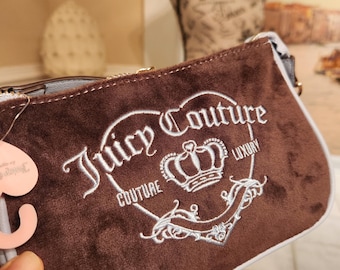 May include: A brown rectangular clutch bag with white embroidered 'Juicy Couture' text and a crown design. It has a light blue trim and a gold zip. The bag is held by a hand.