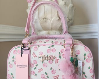Op de afbeelding: Een witte handtas met een roze kersenpatroon en de tekst Juicy Couture. De tas heeft roze afwerkingen, twee in plastic verpakte handvatten, een afneembare schouderriem, een goudkleurig logo, een roze pomponhanger en een papieren label.