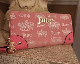 Può includere: Portafoglio rosa con motivo a corona e la scritta "Juicy" in lettere dorate. Il portafoglio ha una cerniera dorata e un ciondolo a forma di cane. Il portafoglio è su una superficie strutturata.