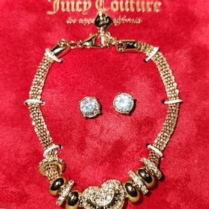 May include: A gold-tone necklace and earrings set with a heart pendant and clear crystal accents. The necklace features a chain with small round beads and the pendant is a large heart with a smaller heart inside. The earrings are small studs with clear crystals.