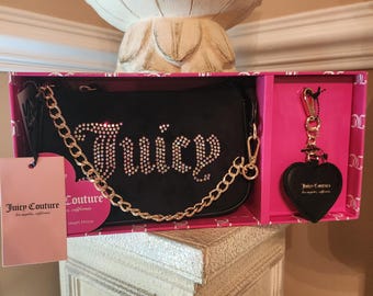 May include: A black Juicy Couture handbag with a gold chain strap and rhinestone embellishments spelling out "Juicy." The bag is presented in a pink box with a matching heart-shaped keychain. A pink tag is attached.