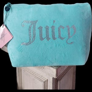 May include: A light blue cosmetic bag with the word "Juicy" spelled out in silver rhinestones. The bag has a zipper closure and a small tag attached. The bag is made of a soft, plush material.