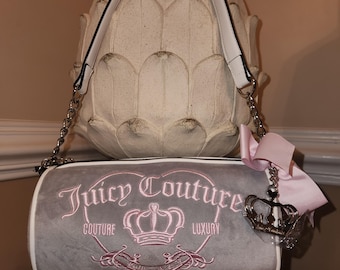 May include: A grey Juicy Couture barrel bag with white trim, a white handle, and silver chain straps. The bag features the brand name and crown logo in pink embroidery. A pink ribbon and crown charm hangs from the side.