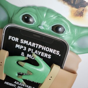 May include: A green Baby Yoda phone holder with a tan arm and hand. The holder is designed to hold smartphones, MP3 players, and more. The text on the holder reads "FOR SMARTPHONES, MP3 PLAYERS & MORE." It has adjustable height, hold, and angles.