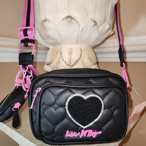 May include: A black quilted crossbody bag with a pink and black strap. The bag has a large heart-shaped patch with rhinestones and the words "Love Betsey" written in pink.