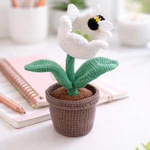 May include: A crocheted white flower with green leaves and a brown pot. A small bee is attached to the flower. The flower is sitting on a white surface next to a pink notebook and a pen. The flower pot is approximately 3 inches tall.
