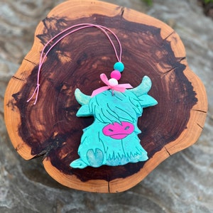 May include: A teal blue highland cow charm with pink and white beads and a pink ribbon. The charm is hanging from a pink string.