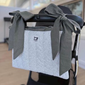 May include: A rectangular pram organiser bag featuring a light grey fabric with a delicate white floral pattern. The bag is secured to a black pram handle with two large, olive green fabric bows. A small white label displays a sewing machine icon.
