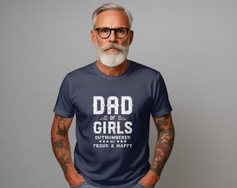 May include: Navy blue t-shirt with the text "DAD OF GIRLS" in a distressed white font. The shirt also includes the phrases "OUTNUMBERED BUT PROUD & HAPPY" and is worn by a man with glasses and a beard.