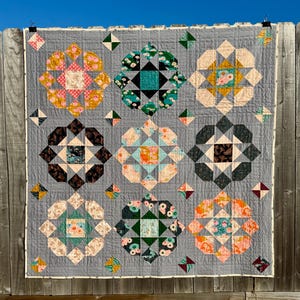 May include: A handmade quilt with a gray background and nine large floral-like blocks in various colors, including pink, teal, and orange. Each block has a central square and is surrounded by geometric shapes. The quilt is hanging on a wooden fence.