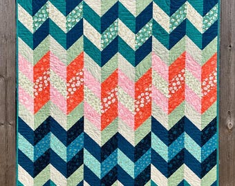 May include: A rectangular quilt featuring a repeating chevron pattern in shades of navy blue, teal, mint green, coral, and soft pink. The fabric includes solid colours and various floral or dotted prints, all finished with a teal binding edge.