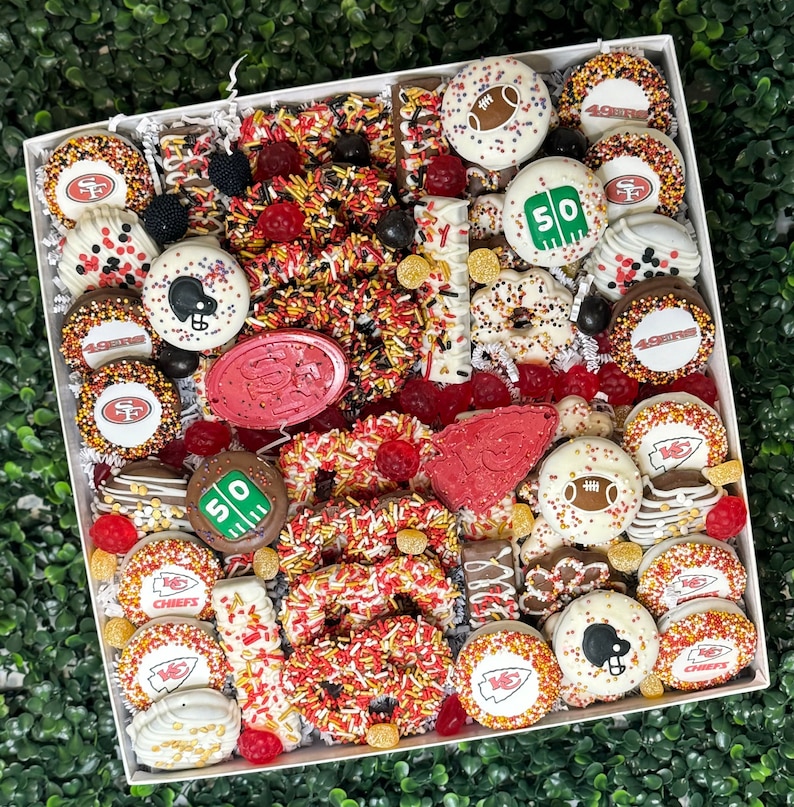 May include: A white box filled with a variety of chocolate treats, including pretzels, candies, and chocolate covered pretzels. The treats are decorated with red, white, and gold sprinkles. The box also contains chocolate footballs, chocolate chips, and chocolate covered strawberries. The treats are arranged in a decorative pattern. The box is a gift basket for a football fan.