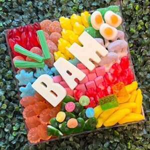 May include: A square clear container filled with an assortment of colorful gummy candies, including red, yellow, orange, green, and blue shapes, topped with large white chocolate letters spelling out the word BAH and green candy sticks.