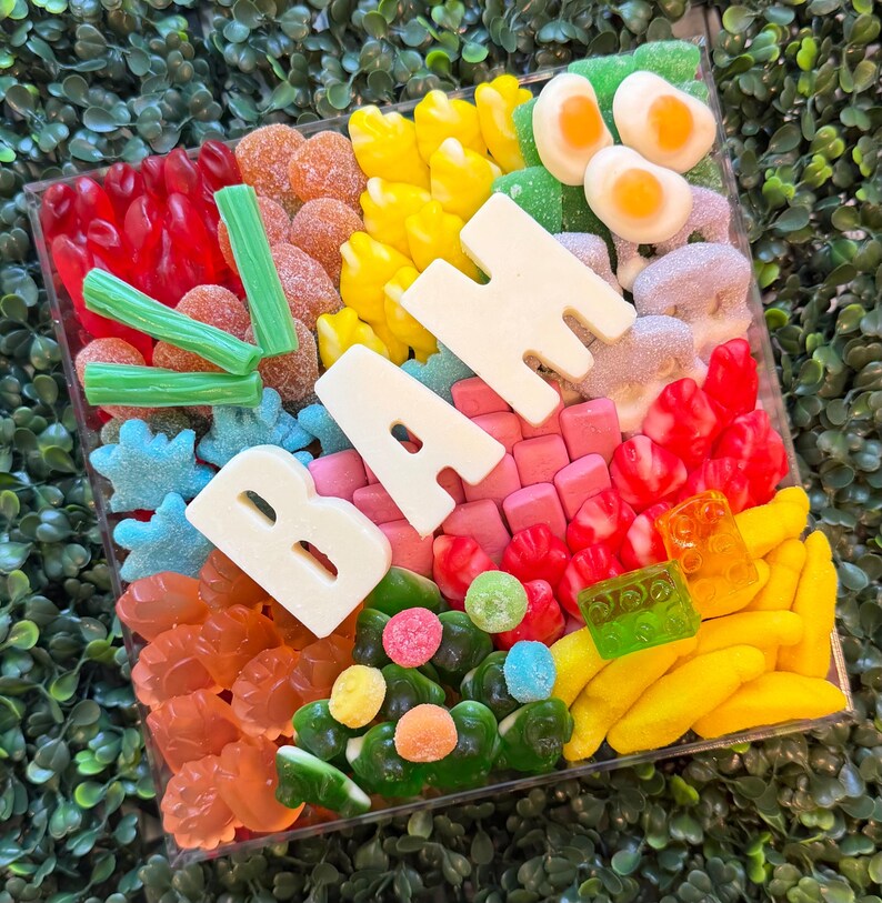 May include: A square clear container filled with an assortment of colorful gummy candies, including red, yellow, orange, green, and blue shapes, topped with large white chocolate letters spelling out the word BAH and green candy sticks.