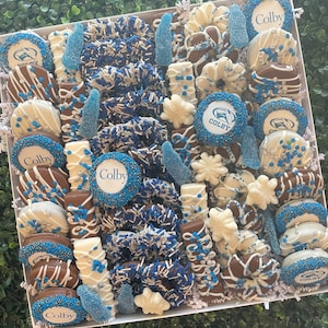 May include: A white box overflowing with an assortment of chocolate-covered treats. The treats include pretzels, cookies, and candies, decorated with blue and white sprinkles and the word "Colby". The arrangement is set against a green backdrop.