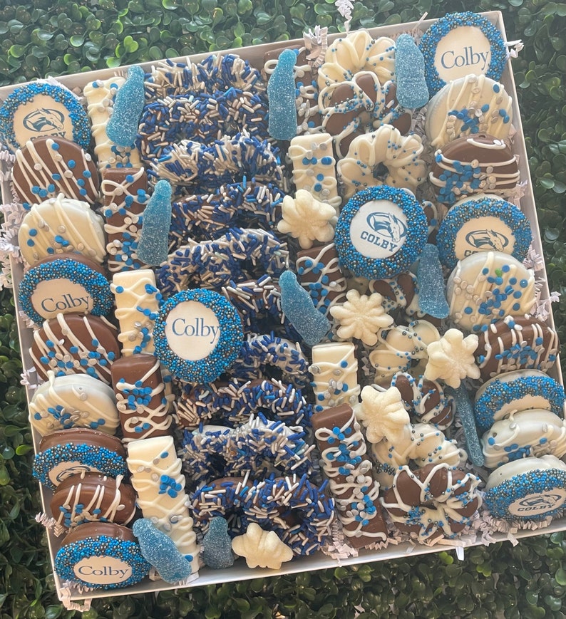 May include: A white box overflowing with an assortment of chocolate-covered treats. The treats include pretzels, cookies, and candies, decorated with blue and white sprinkles and the word "Colby". The arrangement is set against a green backdrop.
