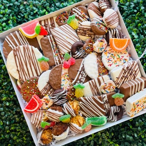 May include: A white box overflowing with an assortment of chocolate-covered treats. The treats include cookies, pretzels, and candies, decorated with sprinkles, drizzled chocolate, and colorful gummy fruit slices. The box is presented against a backdrop of green foliage.