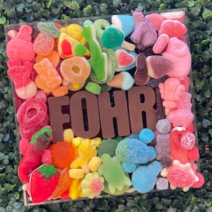 May include: A square clear container filled with a variety of colorful gummy candies in shapes including strawberries, turtles, and unicorns, surrounding large brown chocolate letters that spell out the word FOHR in the center of the arrangement.