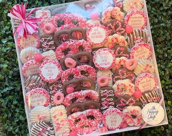 May include: A square gift box filled with chocolate-covered pretzels, cookies, and circular treats decorated with pink and white sprinkles. Several items feature printed text including KICK CANCER, SURVIVED THE CHOP, and FIGHT FLAT WIN BIG.