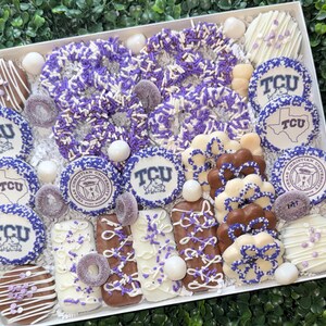 May include: A white box filled with an assortment of decorated cookies, pretzels, and candies. The treats are decorated in purple and white, with some featuring the TCU logo and the words "Christian University".