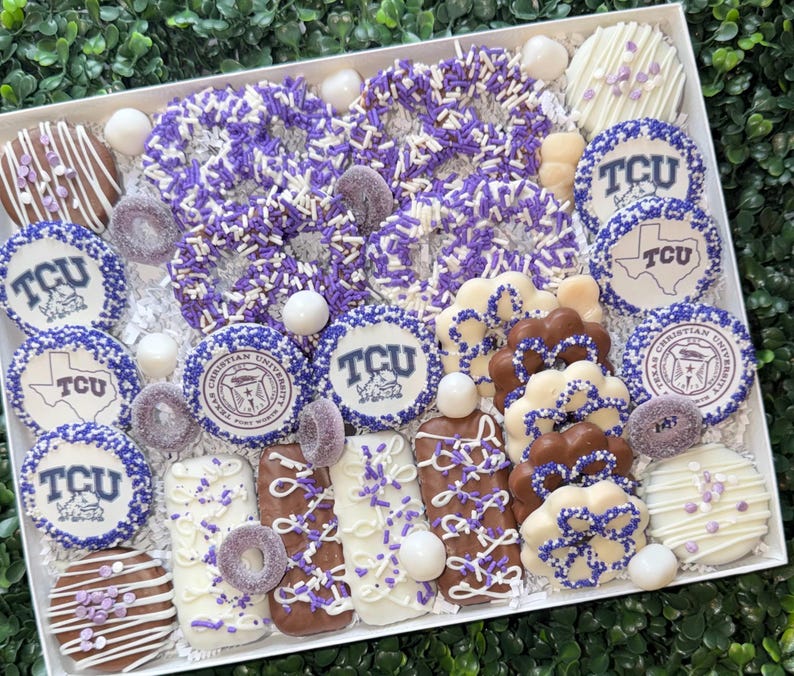 May include: A white box filled with an assortment of decorated cookies, pretzels, and candies. The treats are decorated in purple and white, with some featuring the TCU logo and the words "Christian University".
