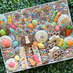 May include: A gift box containing a selection of chocolate-covered Easter treats. The box includes pretzels, biscuits, and bars, all decorated with colourful sprinkles, sweets, and Easter-themed decorations. The treats are arranged on a bed of white paper.