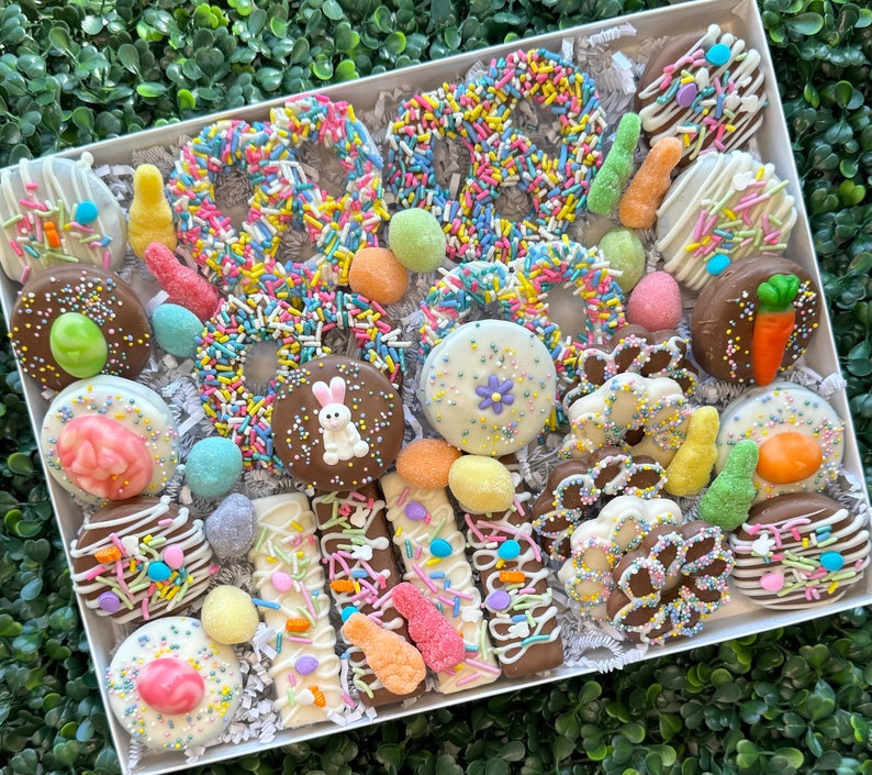 May include: A gift box containing a selection of chocolate-covered Easter treats. The box includes pretzels, biscuits, and bars, all decorated with colourful sprinkles, sweets, and Easter-themed decorations. The treats are arranged on a bed of white paper.