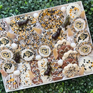 May include: A white rectangular box filled with an assortment of decorated treats. The treats include cookies with a university logo, pretzels with sprinkles, and chocolate-covered items. The color scheme is black, gold, and white.